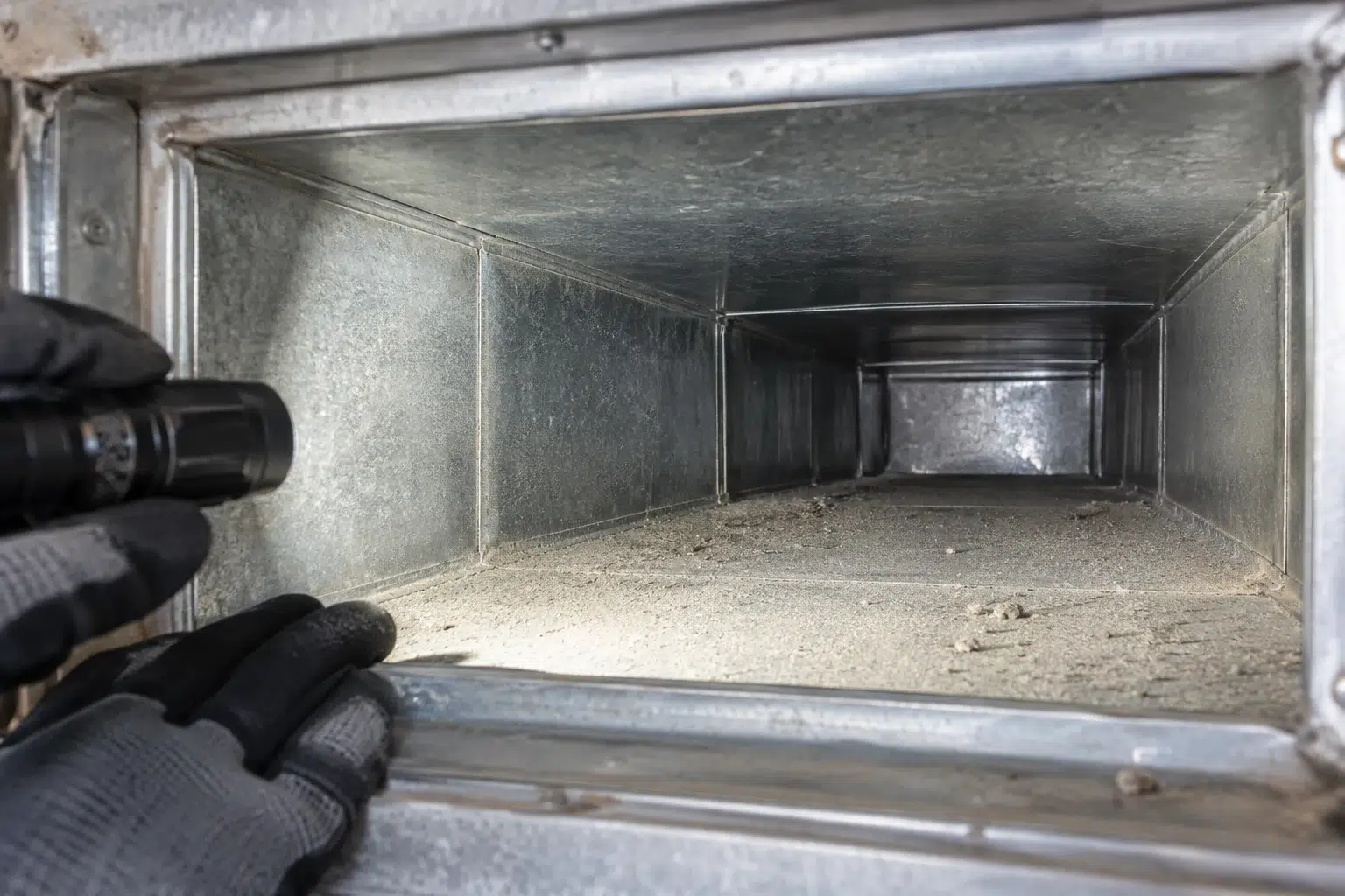 Gloved hand illuminating a light dust deposit inside an accessible metal office duct